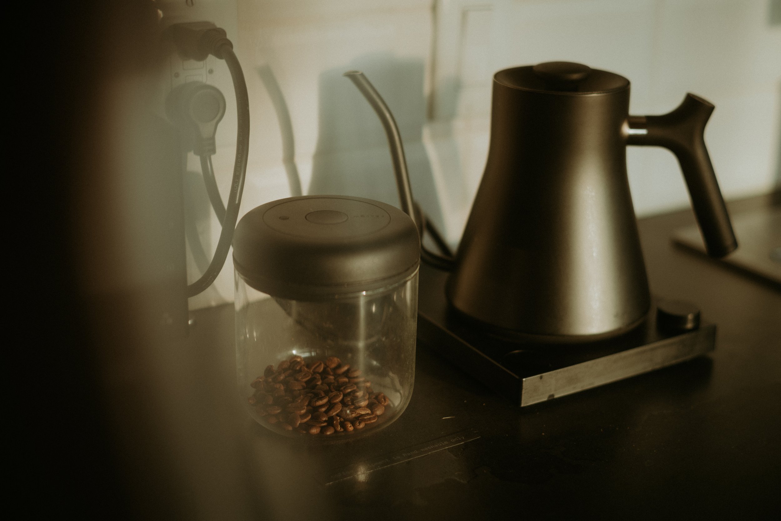 A glass jar filled with coffee beans, a black electric kettle, and a power outlet with cords on a dark kitchen countertop.