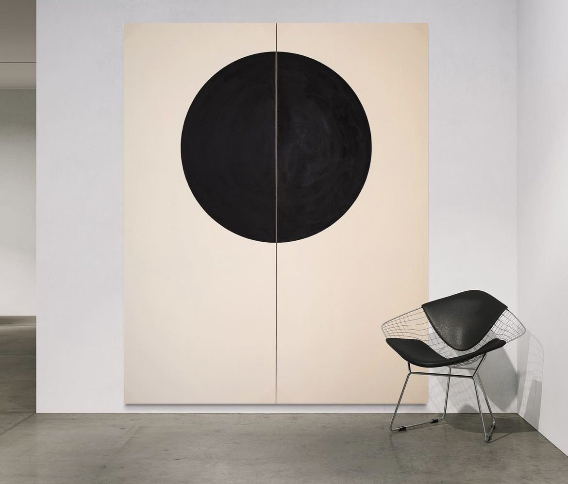a black circle artwork on a beige divided canvas behind a black wire-frame chair